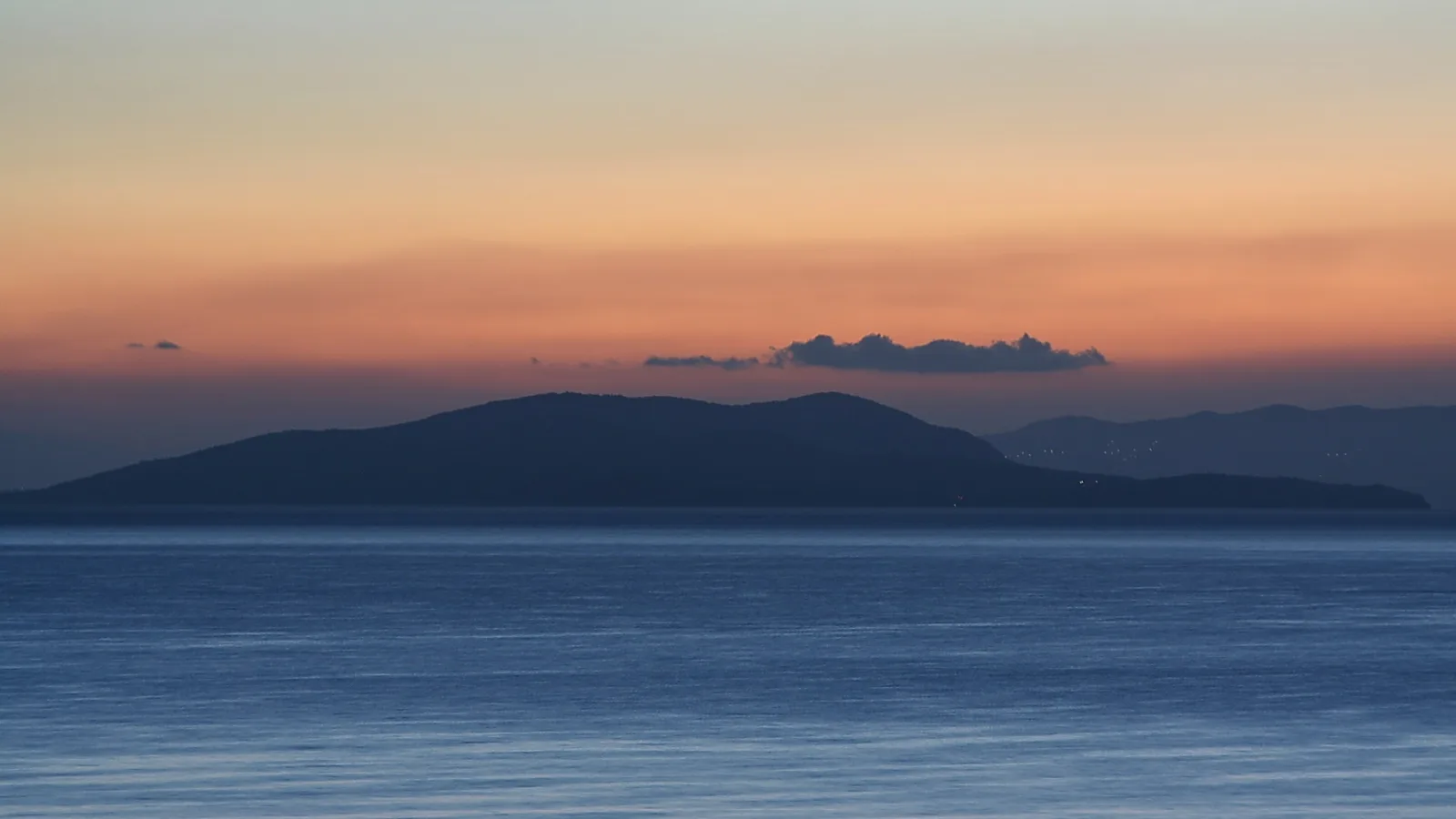 Dunkle Silhouette einer Insel vor einem orange-blau gefärbten Abendhimmel, davor ruhige blaue Meeresfläche