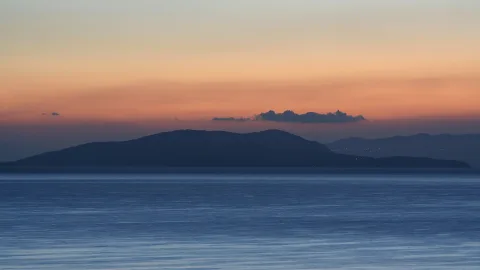 Dunkle Silhouette einer Insel vor einem orange-blau gefärbten Abendhimmel, davor ruhige blaue Meeresfläche