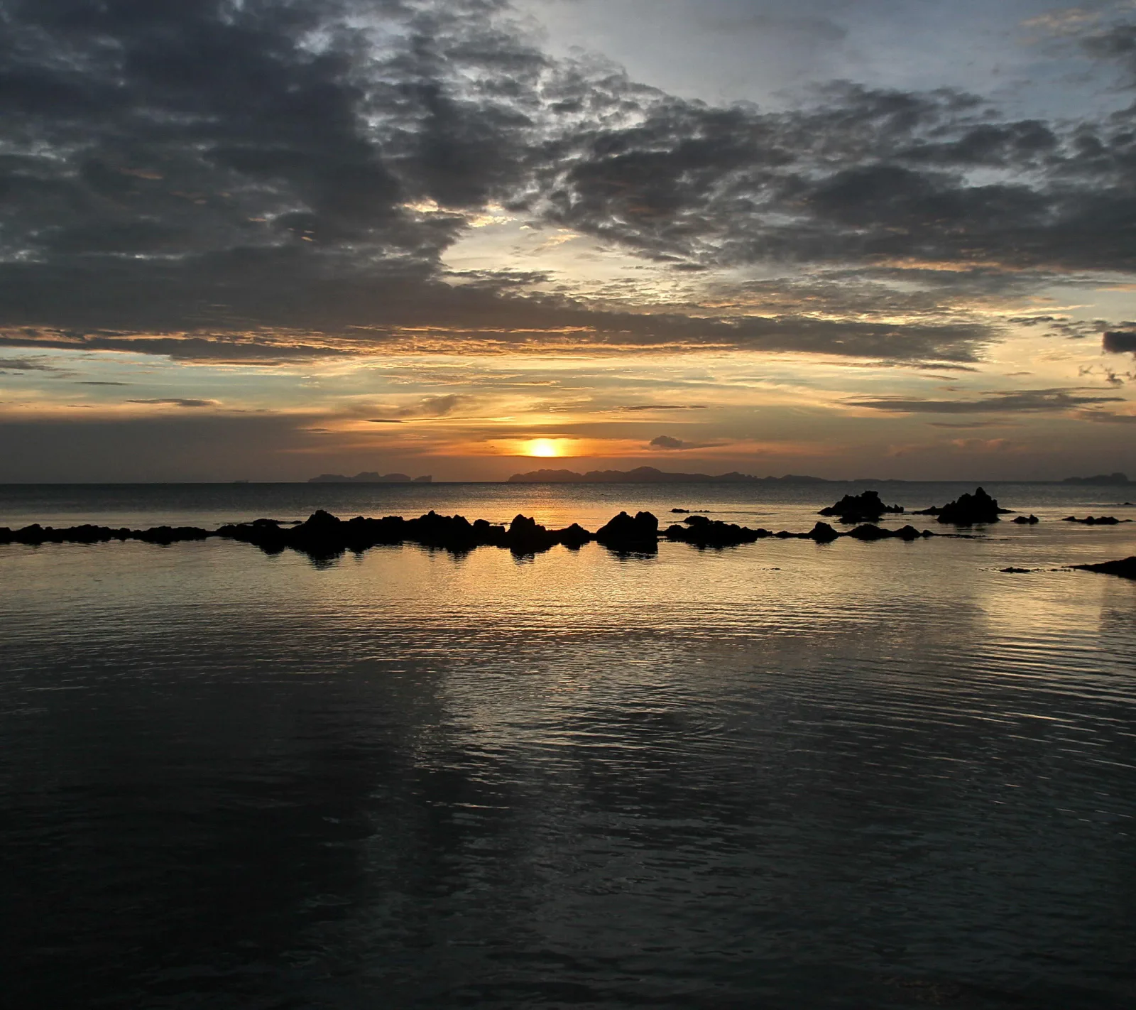 Sonnenuntergang über dem Meer, dunkle Felsen ragen aus dem Wasser, darüber ein Band aus Wolken und fernen Inseln am Horizont.