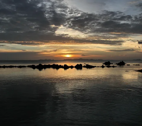 Sonnenuntergang über dem Meer, dunkle Felsen ragen aus dem Wasser, darüber ein Band aus Wolken und fernen Inseln am Horizont.