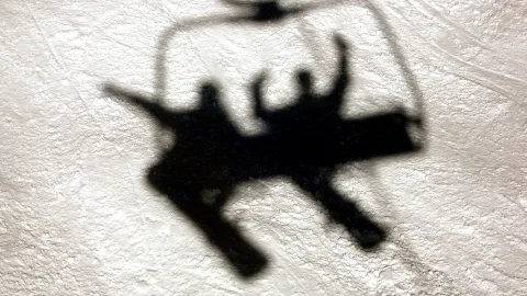 Schwarzer Schattenriss eines Sessellifts mit zwei Personen auf hellem, körnigem Schnee.