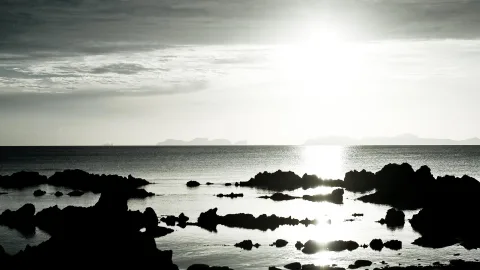 Schwarze Felsen ragen aus einem flachen Meer, im Hintergrund strahlt die Sonne durch einen grauen Himmel, am Horizont liegen Inseln und Berge.
