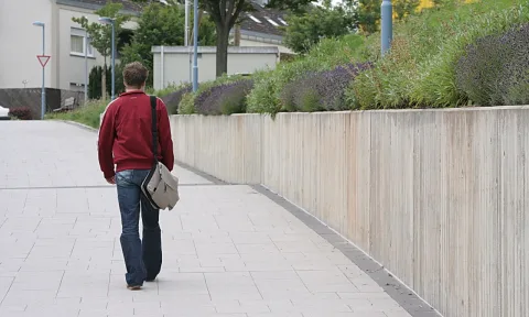 Ein Mann in roter Jacke und Jeans geht von hinten über einen hellen Gehweg neben einer Betonmauer mit blühendem Lavendel. Im Hintergrund stehen Laternen und Wohnhäuser.