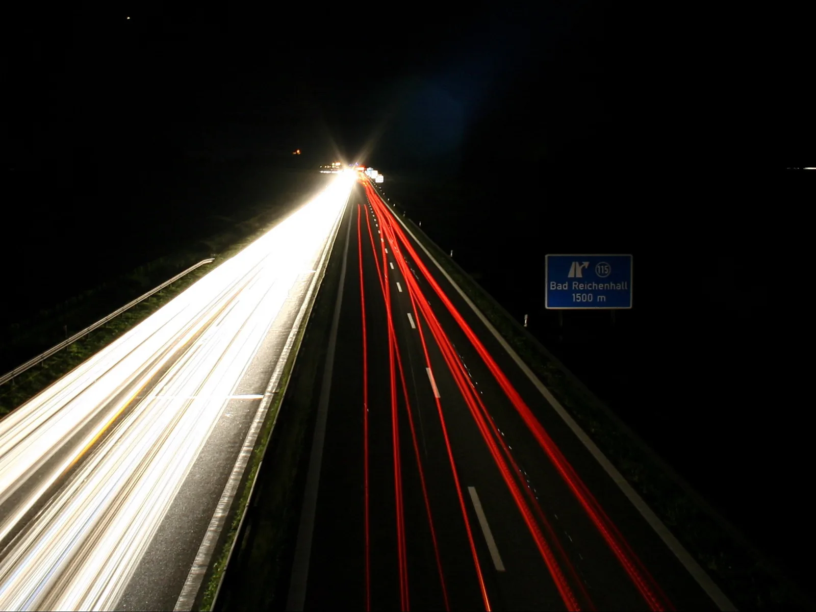 Langzeitaufnahme einer nächtlichen Autobahn mit weißen Scheinwerferspuren links und roten Rücklichtspuren rechts, im Hintergrund ein blaues Schild mit der Aufschrift Bad Reichenhall 1500 m.