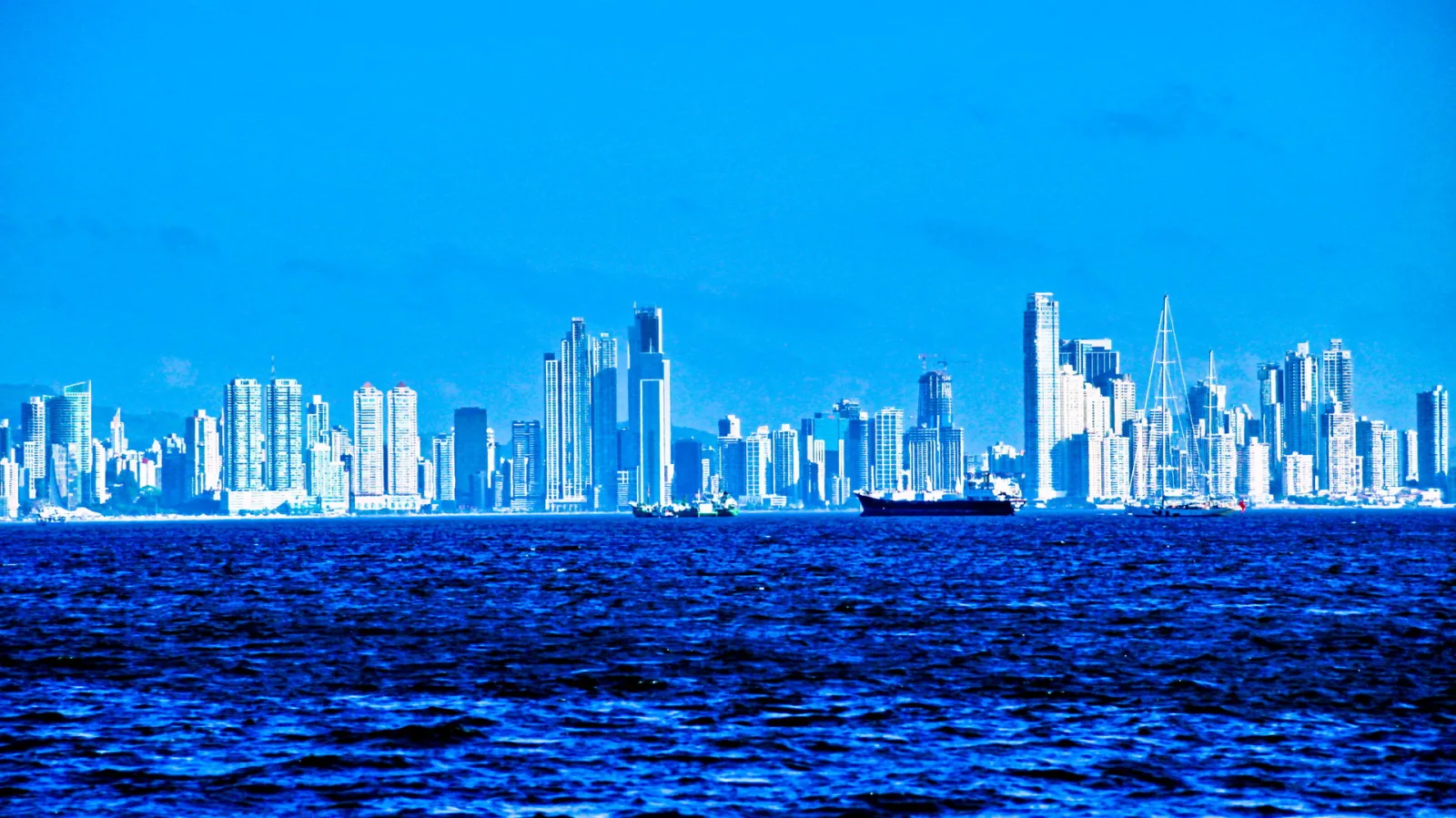 Hochhäuser der Skyline von Panama City entlang der Küste, davor ein dunkles Frachtschiff auf blauem, leicht bewegtem Meer unter wolkenlosem Himmel.