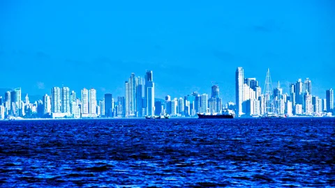 Hochhäuser der Skyline von Panama City entlang der Küste, davor ein dunkles Frachtschiff auf blauem, leicht bewegtem Meer unter wolkenlosem Himmel.