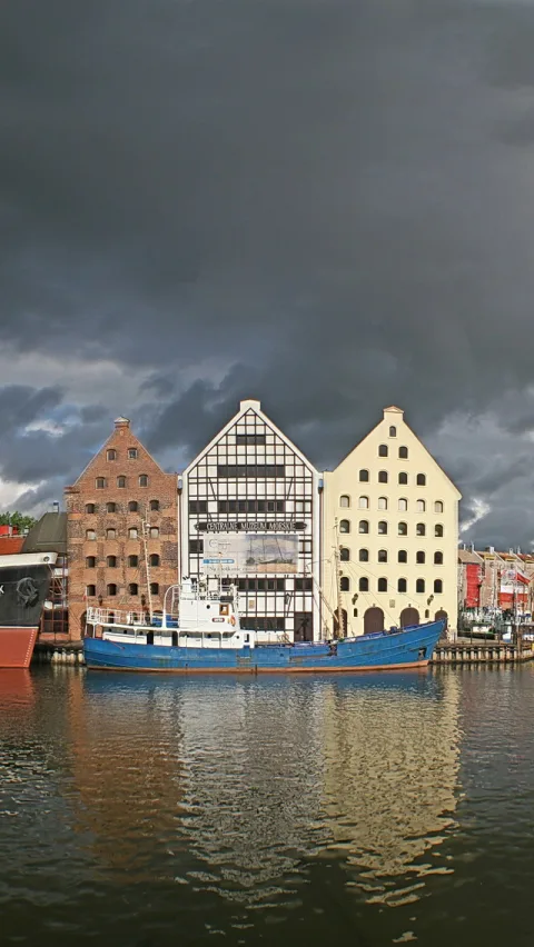 Drei historische Speicherhäuser mit Giebeln stehen am Ufer, davor liegt ein blaues Schiff im Wasser, darüber dunkle Gewitterwolken.