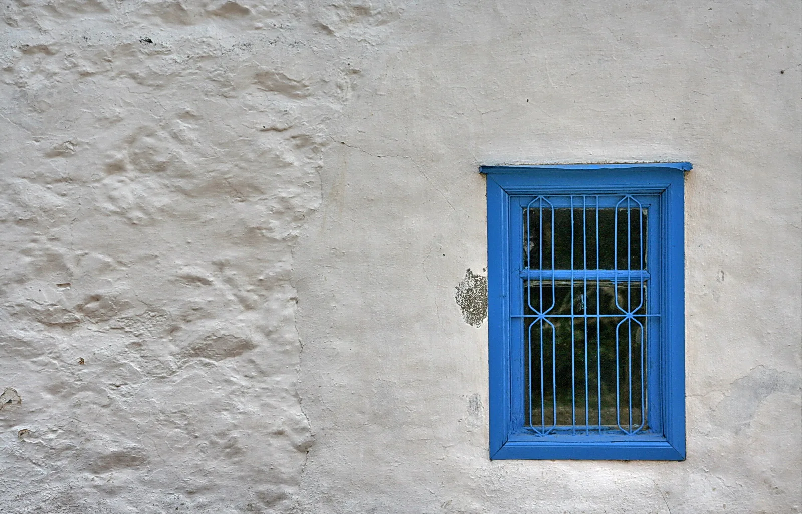 Weiße, unebene Hauswand mit einem kleinen Fenster, dessen blauer Rahmen und blaue Gitterstäbe auffallen. Dahinter ist dunkles Grün zu erkennen.