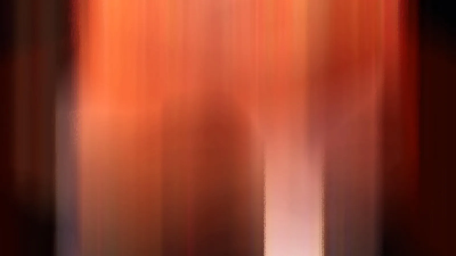 Unscharfe, senkrechte Schlieren in Orange und Rot, in der Mitte unten ein helles rosafarbenes Licht, dunkle Ränder links und rechts.