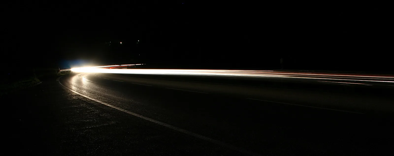 Langzeitbelichtung einer kurvigen Straße bei Nacht: Scheinwerferlicht zieht als helle Spur in die Ferne, rote Rücklichter schimmern am rechten Rand, der Rest liegt im Dunkeln.