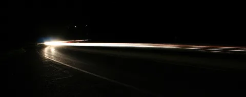 Langzeitbelichtung einer kurvigen Straße bei Nacht: Scheinwerferlicht zieht als helle Spur in die Ferne, rote Rücklichter schimmern am rechten Rand, der Rest liegt im Dunkeln.