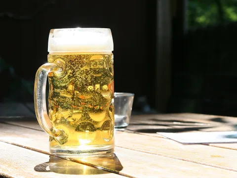 Ein Bierkrug mit goldgelbem Helles und weißer Schaumkrone steht auf einem Holztisch, von der Sonne beschienen. Rechts daneben ein kleines Glas, der Hintergrund ist dunkel.