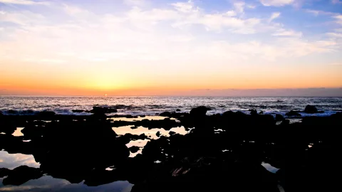 Dunkle Felsen und Gezeitentümpel im Vordergrund, dahinter das Meer und ein orange glühender Horizont bei Sonnenuntergang.