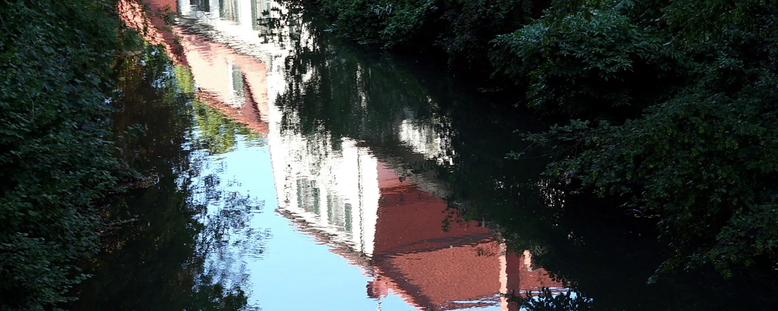 Weiße Hausfassaden mit roten Dächern spiegeln sich im ruhigen Wasser eines Flusses, gesäumt von dichtem grünem Laub.