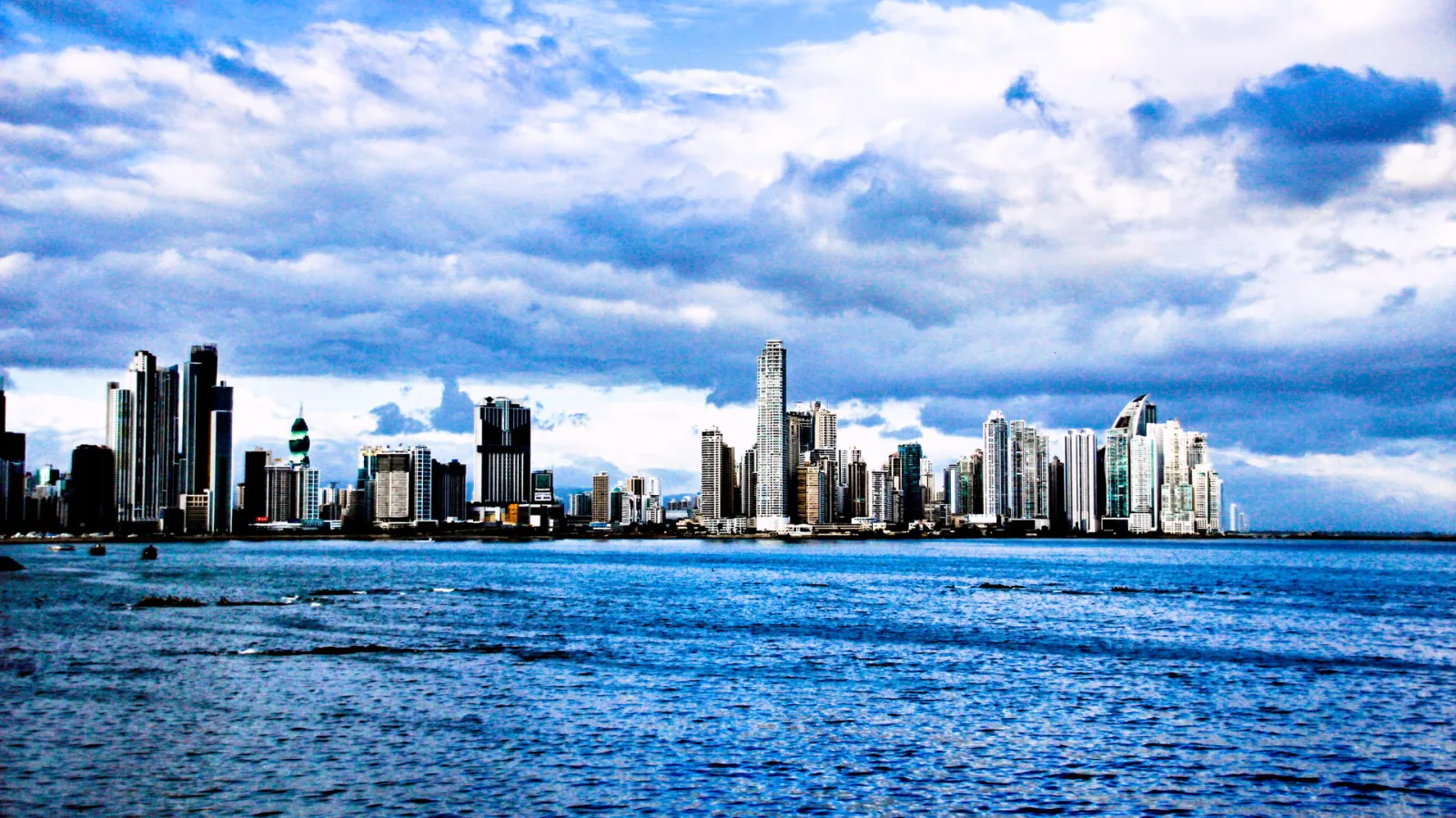Skyline von Panama-Stadt mit dicht stehenden Hochhäusern hinter einer blauen Wasserfläche unter dramatischer Wolkendecke