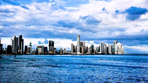 Skyline von Panama-Stadt mit dicht stehenden Hochhäusern hinter einer blauen Wasserfläche unter dramatischer Wolkendecke
