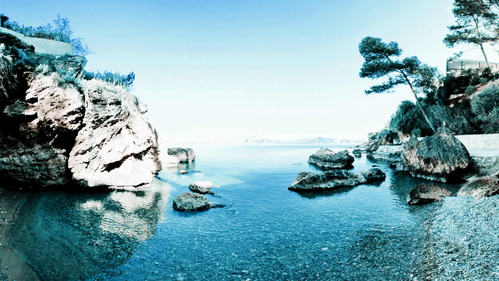Felsige Bucht mit klarem blauem Wasser, im Vordergrund Felsbrocken, rechts eine schräg stehende Kiefer und Büsche am Ufer, im Hintergrund heller Horizont.