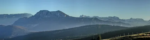 Breites Panorama mit einem schneebedeckten Bergrücken in der Mitte, davor bewaldete Hänge und Dunst in den Tälern, darüber klarer hellblauer Himmel.