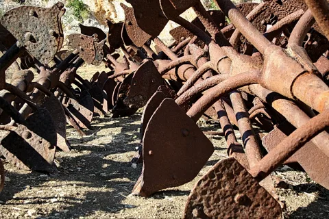 Stapel verrosteter landwirtschaftlicher Metallteile mit runden Scheiben und Stangen auf sandigem Boden, im Hintergrund heller Fels.