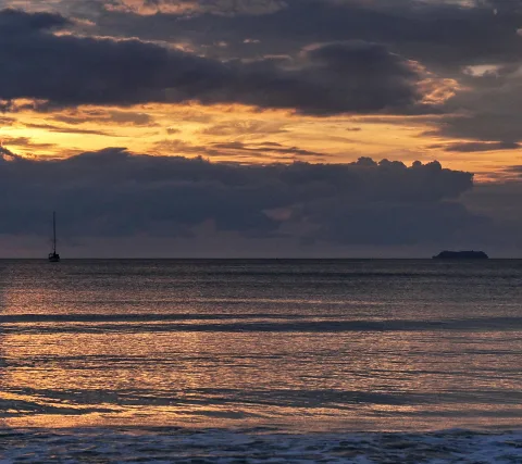 Ein kleines Segelboot links und eine dunkle Insel rechts am Horizont, darüber dunkle Wolken und oranger Himmel bei Sonnenuntergang über dem Meer.