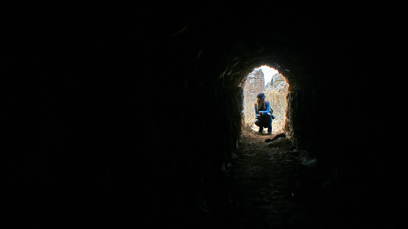 Eine Person in dunkler Jacke hockt im Ausgang eines schmalen Steintunnels, dahinter sind helle Felsen und Mauern zu sehen.