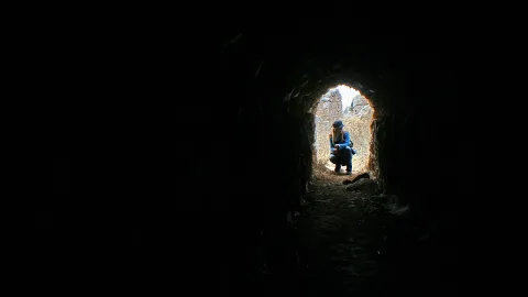Eine Person in dunkler Jacke hockt im Ausgang eines schmalen Steintunnels, dahinter sind helle Felsen und Mauern zu sehen.