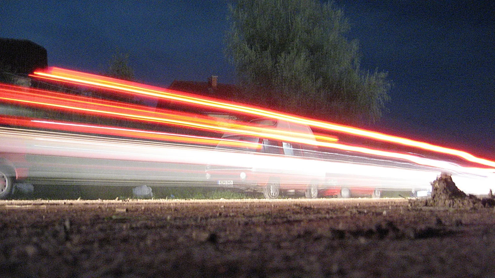Langzeitbelichtung einer nächtlichen Straße mit roten Rücklichtspuren und weißen Lichtstreifen über einem parkenden Fahrzeug, im Hintergrund ein Baum und dunkler Himmel.