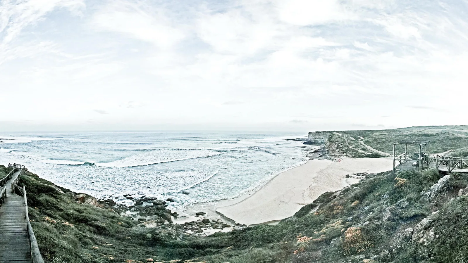 Breites Panorama einer Küste mit Wellen, die auf einen Sandstrand auslaufen. Links führt ein Holzsteg einen grasbewachsenen Hang hinab, rechts steht ein Holzgeländer auf einer Klippe. Der Himmel ist hell und bewölkt.
