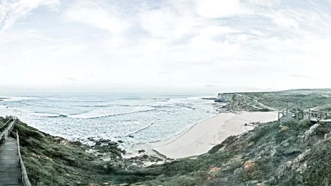 Breites Panorama einer Küste mit Wellen, die auf einen Sandstrand auslaufen. Links führt ein Holzsteg einen grasbewachsenen Hang hinab, rechts steht ein Holzgeländer auf einer Klippe. Der Himmel ist hell und bewölkt.