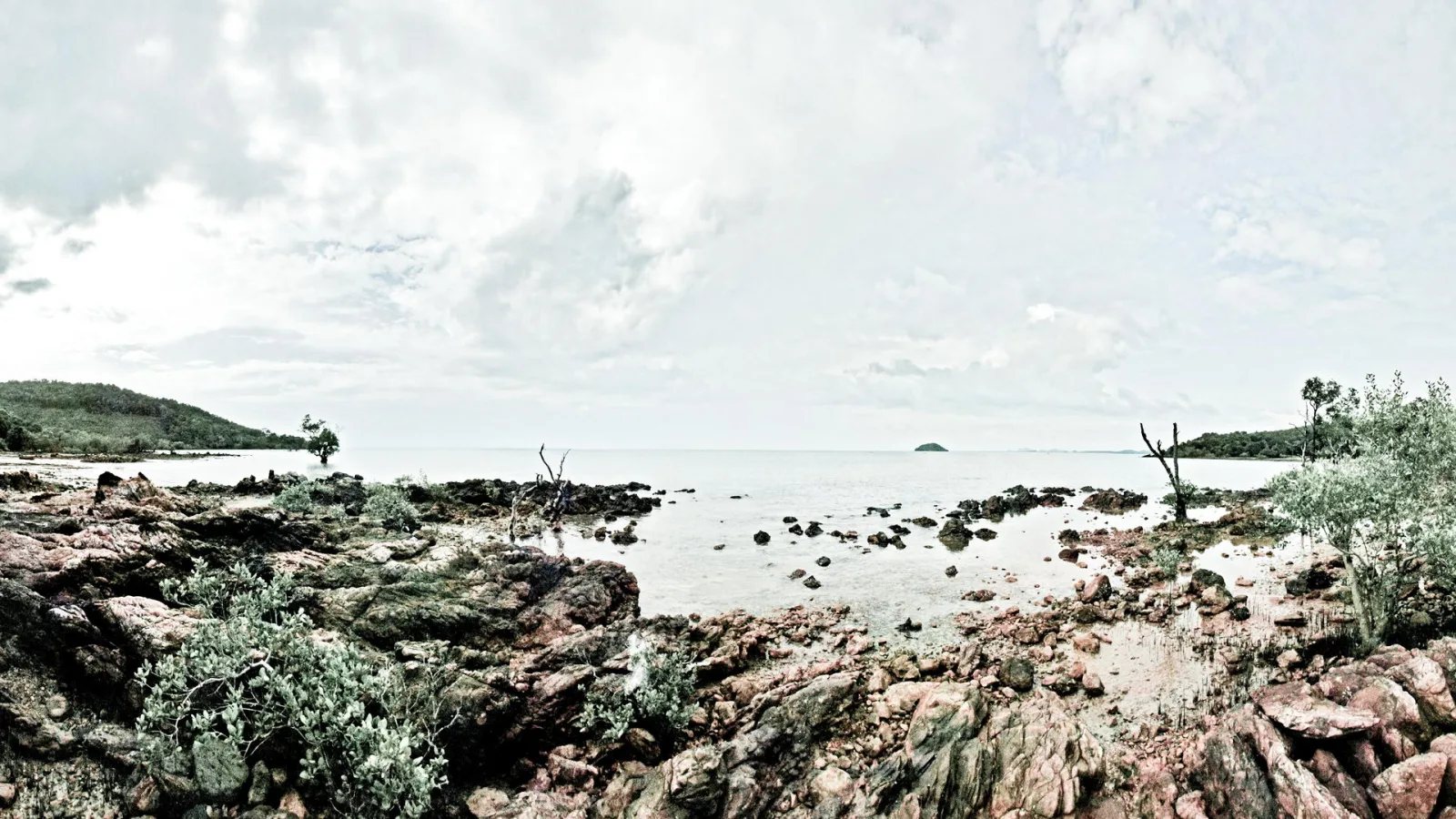 Felsige Küste mit rotbraunen Steinen, flachem Wasser und grünen Büschen, dahinter ein Meer unter bewölktem Himmel und eine kleine Insel am Horizont.
