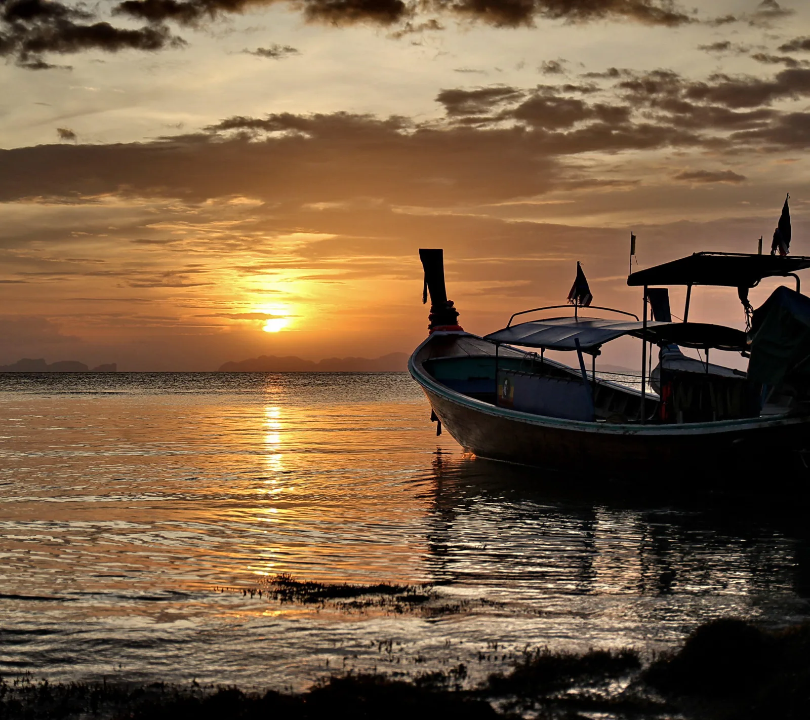 Silhouette eines Longtailbootes mit Dach liegt bei Sonnenuntergang am ruhigen Wasser, im Hintergrund sind flache Inseln zu sehen.