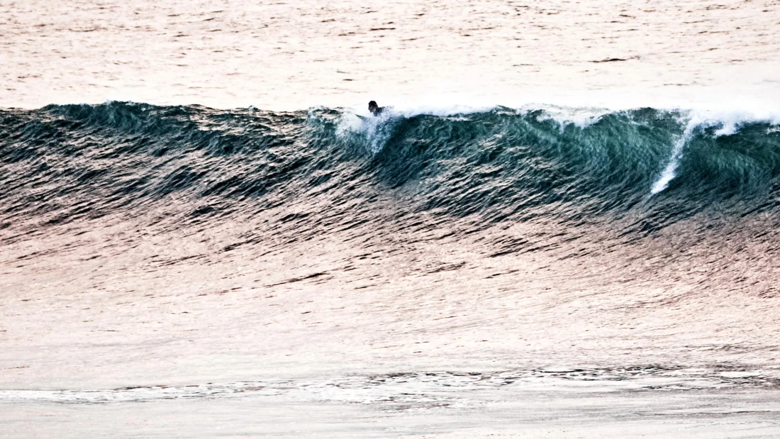 Ein kleiner Surfer gleitet auf einer grünblauen Welle, das Meer schimmert rosa im Abendlicht.