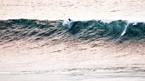 Ein kleiner Surfer gleitet auf einer grünblauen Welle, das Meer schimmert rosa im Abendlicht.