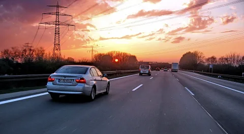 Silberner BMW fährt bei Sonnenuntergang auf einer Autobahn, im Hintergrund Strommasten und Wolken im Abendrot.