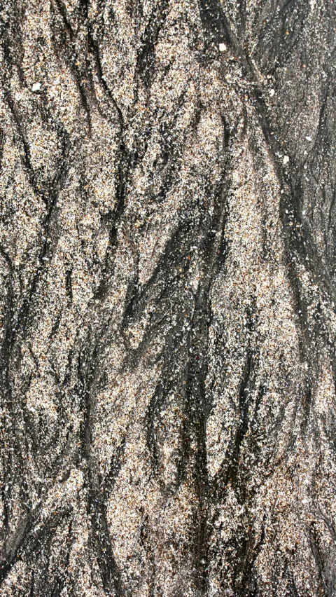 Nahaufnahme einer unebenen Oberfläche aus dunklem Sand mit hellen Körnern und welligen, dunklen Rillen, die von oben nach unten verlaufen.