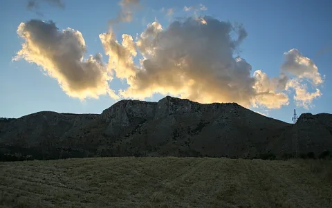Felsiger Bergrücken unter einer Wolkenbank, davor eine trockene Grasfläche und ein Strommast am rechten Bildrand.
