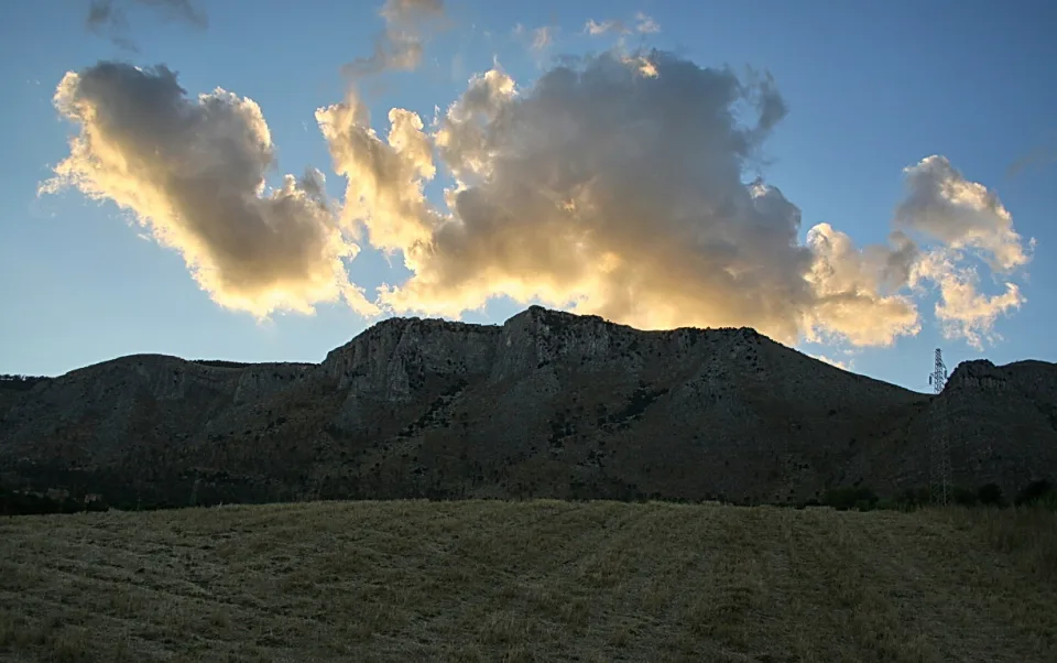 Felsiger Bergrücken unter einer Wolkenbank, davor eine trockene Grasfläche und ein Strommast am rechten Bildrand.