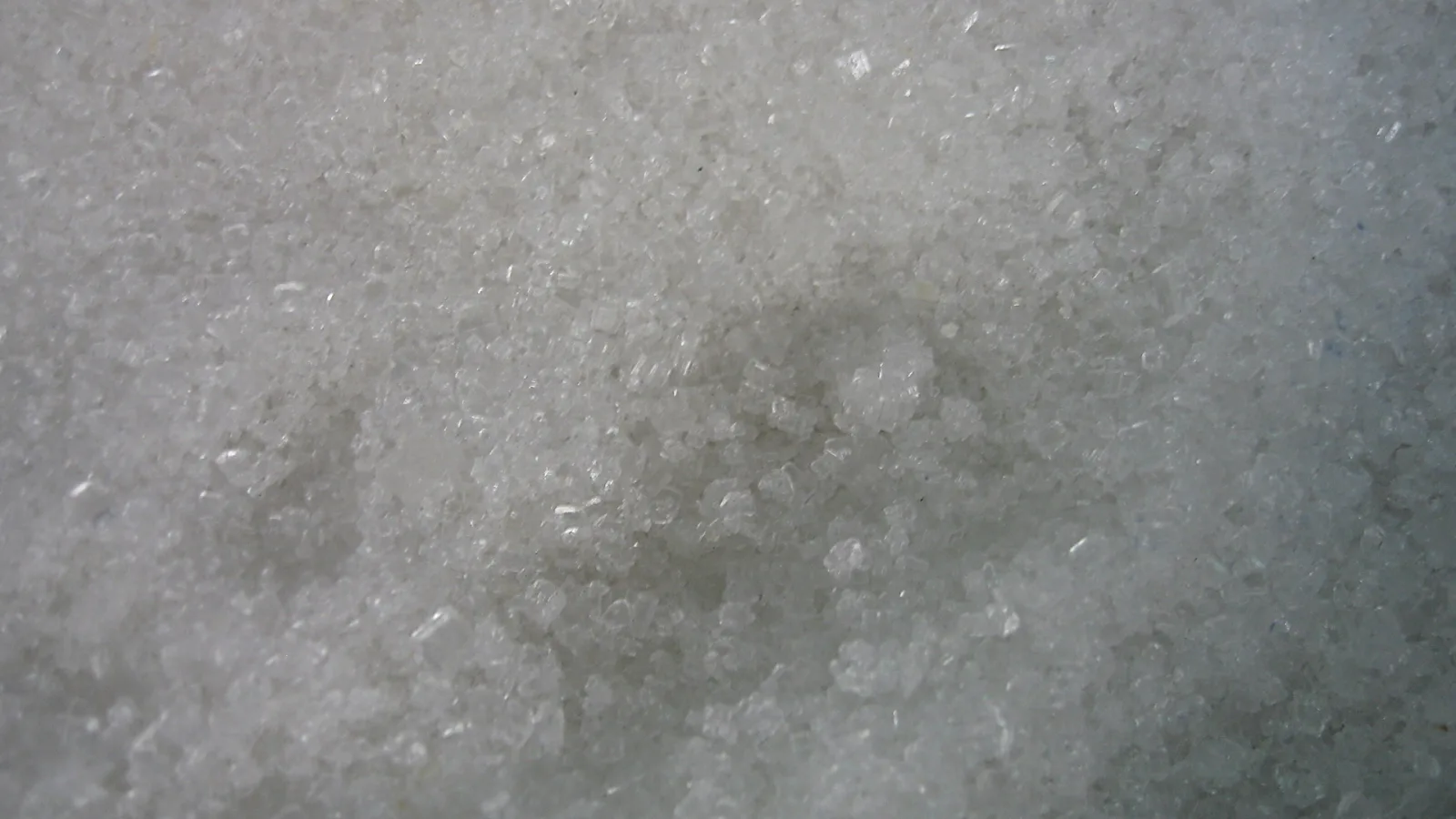 Nahaufnahme von dicht gepackten, weißen Kristallkörnern mit glitzernden Kanten und Lichtreflexen.
