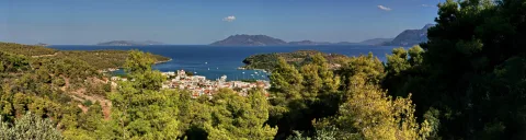 Panorama einer kleinen Küstenstadt mit Hafen und vielen Booten, umgeben von Pinienwald, davor blaues Meer und dahinter Bergketten unter klarem Himmel.