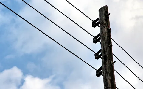 Ein hoher Holzmast mit Porzellanisolatoren, von dem mehrere Stromleitungen diagonal ins Bild laufen, vor einem blau-weiß bewölkten Himmel.