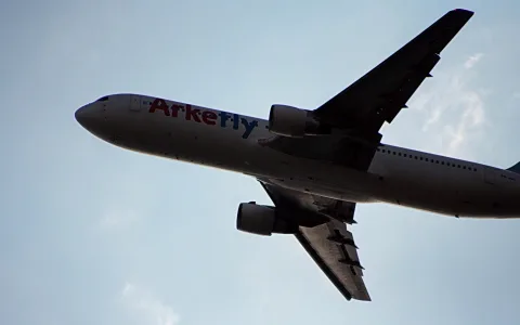 Passagierflugzeug der Airline Arkefly mit roter und blauer Schrift am Rumpf fliegt schräg von unten gesehen vor hellblauem Himmel.