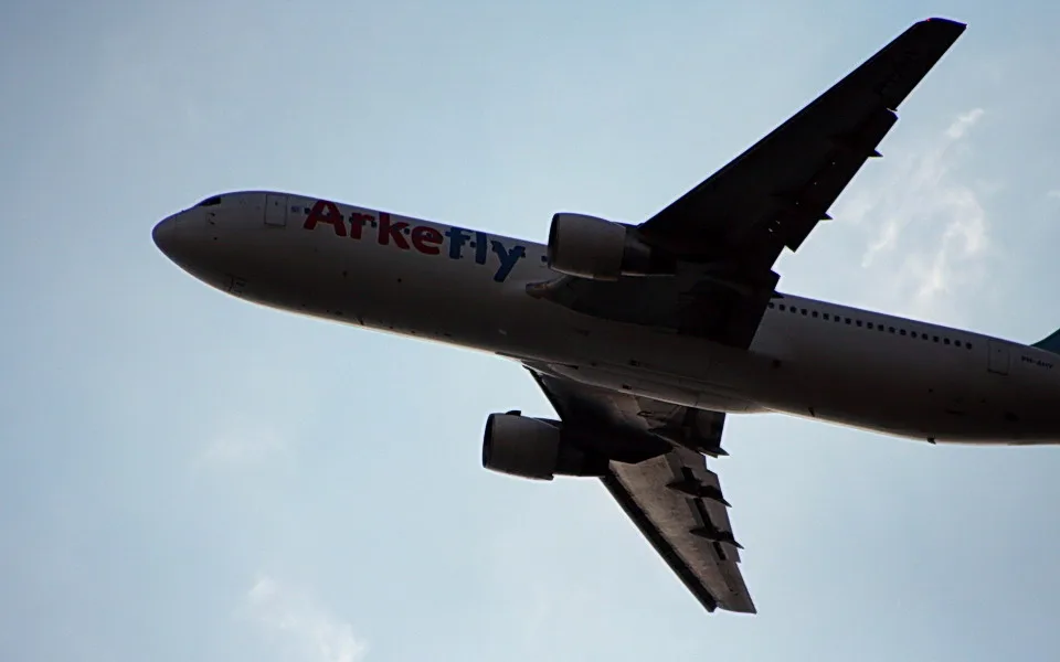 Passagierflugzeug der Airline Arkefly mit roter und blauer Schrift am Rumpf fliegt schräg von unten gesehen vor hellblauem Himmel.