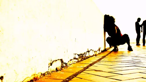 Hockende Person als dunkle Silhouette vor einer hellen Mauer, rechts zwei stehende Personen auf gelb leuchtendem Pflaster