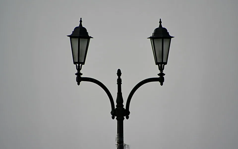 Schwarze Straßenlaterne mit zwei Laternen und verzierten Armen vor grauem Himmel, symmetrisch aufgebaut.