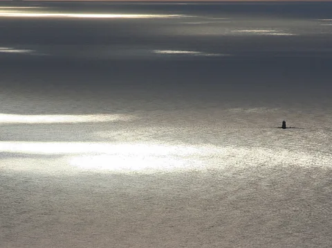 Weite Meeresfläche mit silbrig glitzernden Lichtreflexen der Sonne, rechts im Bild eine kleine dunkle Turmsilhouette auf dem Wasser.