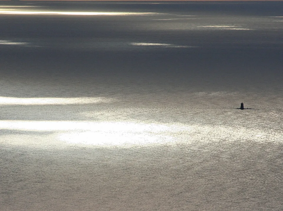 Weite Meeresfläche mit silbrig glitzernden Lichtreflexen der Sonne, rechts im Bild eine kleine dunkle Turmsilhouette auf dem Wasser.
