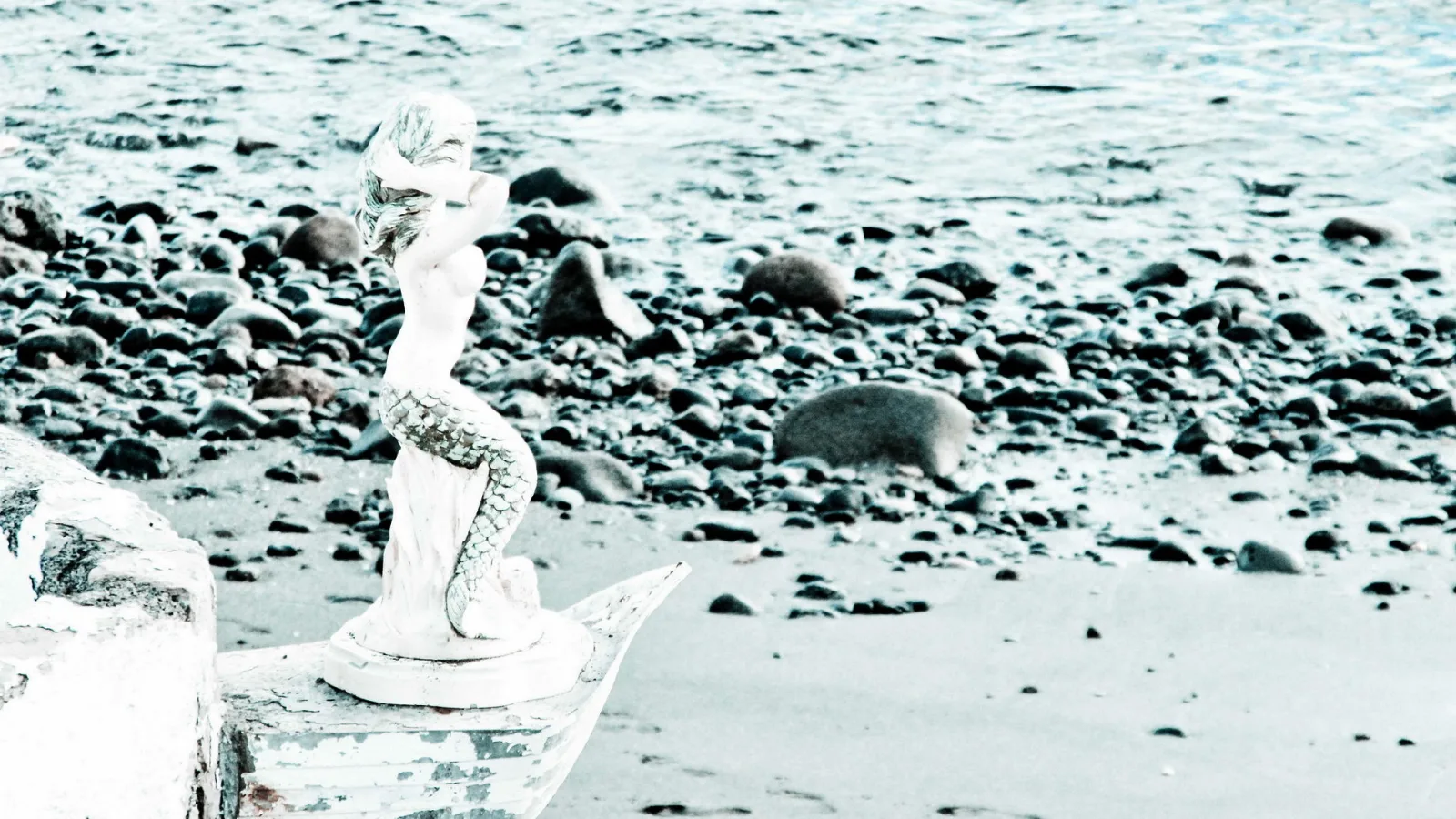 Weiß getönte Meerjungfrau-Statue auf einem Holzboot an einem Kiesstrand, im Hintergrund das Meer.