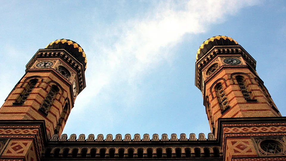 Zwei symmetrische Backsteintürme mit goldverzierten dunklen Zwiebelkuppeln und Turmuhren, aufgenommen von unten vor blauem Himmel mit Wolkenstreifen.