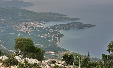 Luftaufnahme eines Küstendorfs mit Olivenhainen, einer Bucht und bewaldeten Bergen am Meer auf der Peloponnes.