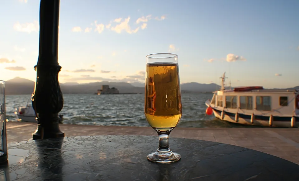 Ein Glas helles Bier auf einem dunklen Marmortisch, im Hintergrund Hafenpromenade mit einem weißen Ausflugsboot und der Festung Bourtzi im Wasser bei Sonnenuntergang.